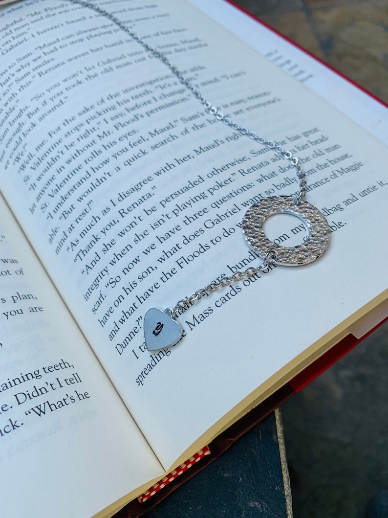 May include: A silver necklace with a heart charm and a round charm with a textured surface. The necklace is lying on a book with text about a fictional character named Maud.