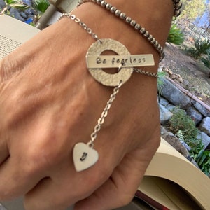 May include: A silver bracelet with a charm that says "Be fearless" and a heart charm with the letter "J" hanging from it.