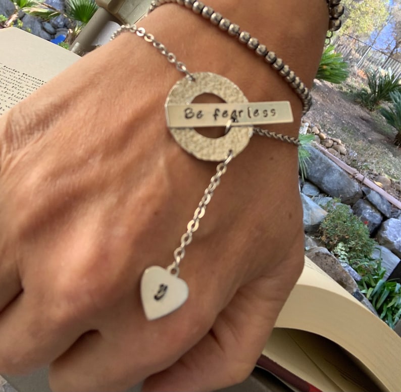 May include: A silver bracelet with a charm that says "Be fearless" and a heart charm with the letter "J" hanging from it.