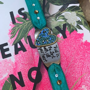 May include: A teal blue elastic bracelet with a silver heart charm. The charm has a blue and green mosaic design and the words "Trust in the Lord with all your heart".