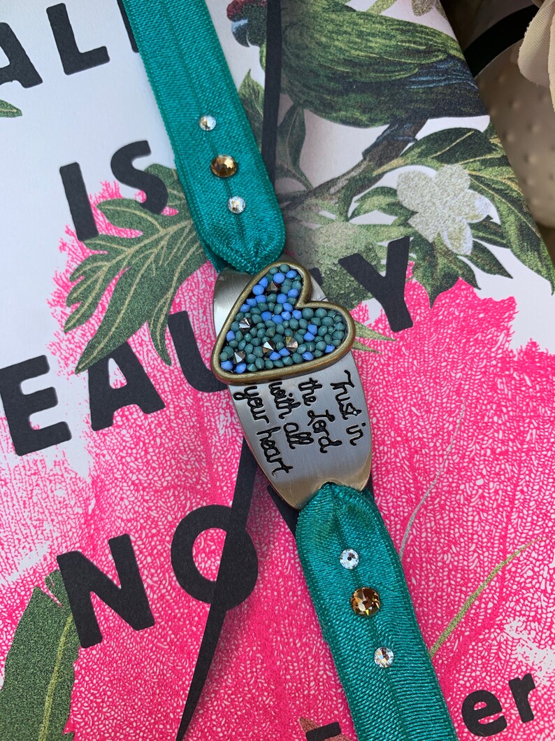 May include: A teal blue elastic bracelet with a silver heart charm. The charm has a blue and green mosaic design and the words "Trust in the Lord with all your heart".