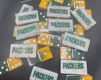 May include: A set of 14 dominoes with a green and white color scheme. Each domino has the word "Packers" printed on it. The dominoes are made of a white material with green glitter.