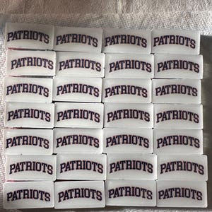 Op de afbeelding: Een verzameling witte, rechthoekige objecten, elk met het woord "PATRIOTS" in marineblauwe, gebogen letters met een rode omtrek. De objecten zijn in nette rijen en kolommen gerangschikt, wat duidt op teammerchandise.