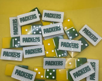 Op de afbeelding: Een verzameling rechthoekige dominostenen met een tweekleurig ontwerp in geel en groen. Elke dominosteen heeft een wit bovengedeelte met het woord PACKERS in donkergroene hoofdletters, terwijl het onderste gedeelte witte stippen bevat.