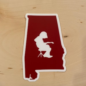 May include: A red outline of the state of Alabama with a white silhouette of a person sitting on a stool inside the outline.