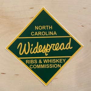 May include: A green diamond-shaped sticker with yellow trim. The sticker says "North Carolina" and "Widespread Ribs & Whiskey Commission" in yellow text.
