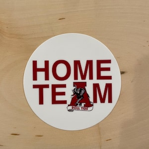 May include: White circle sticker with red text that says "HOME TEAM" with a red Alabama Crimson Tide logo in the center. The logo features an elephant and the words "ROLL TIDE".