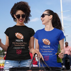 May include: Two women wearing black and blue t-shirts with a football graphic and the text "MOM YOU Discovered Your Favorite Player I RAISED MINE 100".