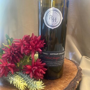 May include: A brown glass candle jar with a black label that reads "Robert Hall Artisan Collection Cabernet Sauvignon". The label also features a silver logo with the initials "RH" and the text "Since 1999". The candle jar is sitting on a wooden surface with red flowers.