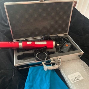 May include: A red device with the text "Quantum Science Energy" is displayed inside a silver case. A black bracelet, blue cloth, and other accessories are also included. The case is open on a black surface.