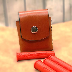 May include: A brown leather pouch with a silver snap closure. Three red cartridges are visible, each labeled "45-70 Government". The pouch and cartridges are set against a light-colored background.