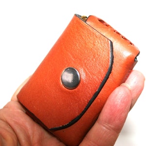 May include: A small, orange leather key case with a silver snap closure. The key case has a curved design with black stitching along the edges. The leather has a textured appearance.