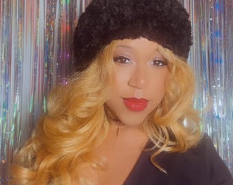 Könnte beinhalten: Eine Person trägt einen schwarzen Kunstpelzhut mit langen, welligen blonden Haaren und rotem Lippenstift. Die Person trägt ein schwarzes Langarm-Top. Der Hintergrund ist ein silberner, glitzernder Vorhang.
