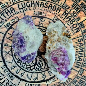 May include: Two pieces of raw amethyst crystal with purple and white hues. The crystals are sitting on a wooden surface with a circular design and text that includes the names of the months and the Celtic Wheel of the Year.