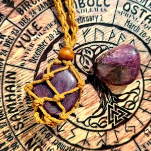 Puede incluir: Un colgante de piedra preciosa púrpura con un diseño de nudo macramé, colgado de un cordón marrón. El colgante está sobre un fondo de madera con un diseño circular y texto que incluye las palabras "Yule", "Ostara" y "Samhain".