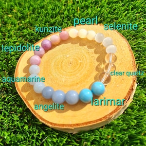Könnte beinhalten: Ein Armband mit Perlen aus verschiedenen Edelsteinen, darunter Aquamarin, Angelit, Larimar und Bergkristall. Das Armband wird auf einer Holzscheibe vor einem Hintergrund aus grünem Gras präsentiert. Die Namen der Steine sind beschriftet.