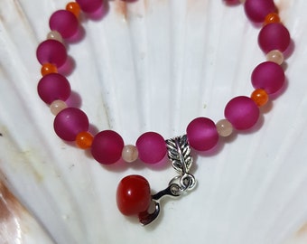 May include: A pink and orange beaded bracelet with a silver charm in the shape of a cherry. The bracelet is on a white seashell background.