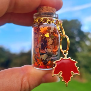 Puede incluir: Un pequeño frasco de vidrio lleno de purpurina colorida, pequeñas piedras y hojas en miniatura. El frasco tiene un tapón de corcho y está unido a una cadena dorada con un dije de hoja de arce roja.