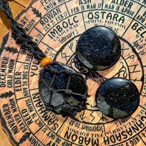 May include: A black obsidian necklace with three round stones, one of which is in a black macrame net bag. The necklace is displayed on a wooden wheel with a calendar of the year, featuring the names of the eight pagan holidays.