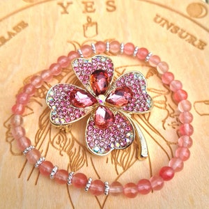 May include: A pink beaded bracelet featuring a gold-coloured four-leaf clover charm. The clover is adorned with pink rhinestones and larger pink gems. The bracelet rests on a wooden surface with the word "YES" visible.