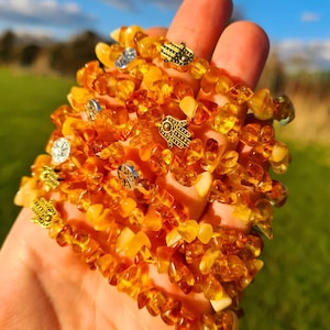 May include: A stack of amber gemstone bracelets with gold and silver charms. The charms are in the shape of a hand with an eye in the centre, known as the Hamsa hand.