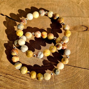 May include: Two bracelets made of yellow, brown, and white agate beads with silver accents. The bracelets are stacked on top of each other on a wooden surface.