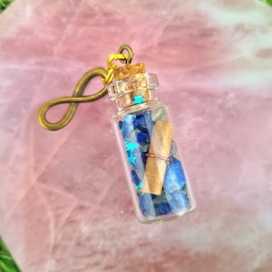 Puede incluir: Una pequeña botella de vidrio con un tapón de corcho y un amuleto de infinito dorado adjunto. La botella está llena de piedras azules y grises y un trozo de papel enrollado. La botella está sobre una superficie rosa.