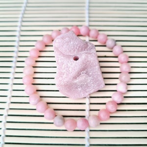 May include: A pink gemstone with a hole in the center, surrounded by a pink beaded bracelet on a light brown bamboo mat.