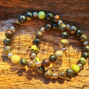 May include: Two bracelets made of green, brown, and yellow gemstone beads with silver accents. The bracelets are on a wooden surface.