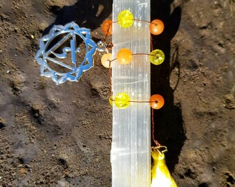 May include: A selenite wand with a beaded tassel and a silver charm featuring a symbol of the sacral chakra. The beads are orange and yellow.