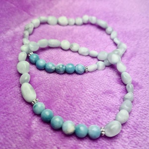 Könnte beinhalten: Zwei Perlenarmbänder, eines mit hellblauen Perlen und das andere mit weißen Perlen. Die Armbänder liegen auf einem violetten Stoffhintergrund.