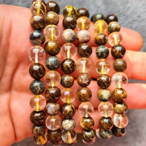 Op de afbeelding: Een set van zes armbanden gemaakt met bruine, gouden en heldere kralen. De kralen zijn rond en hebben een gevlekt uiterlijk.