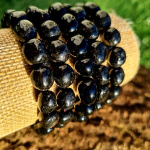 May include: A stack of black beaded bracelets made of polished black obsidian stones. The bracelets are stacked on a beige fabric surface.