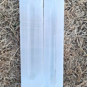 Puede incluir: Dos placas de carga de selenita blanca, una más grande que la otra, están sobre un lecho de hierba seca. Las placas son lisas y tienen una textura estriada sutil.