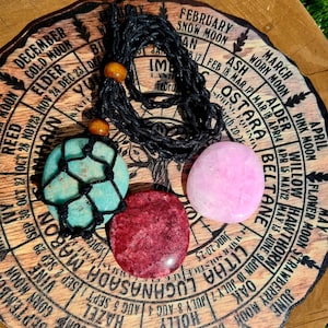 May include: A wooden wheel with a calendar of moon phases and names for each month. Three smooth, flat, round stones in shades of pink, turquoise, and red are arranged on the wheel. A black cord with wooden beads is draped over the stones.