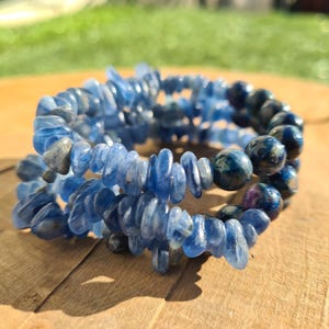 May include: A set of blue gemstone bracelets. The bracelets are composed of small, irregular blue stones and larger, round, speckled blue beads. The bracelets are on a wooden surface, with a blurred green background.