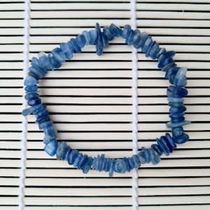 May include: A blue gemstone bracelet made of small, irregular shaped chips. The bracelet is laid out on a white and brown bamboo mat.