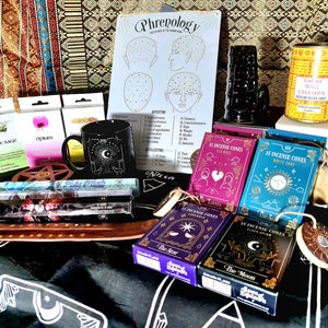 Può includere: Una collezione di articoli spirituali e metafisici, tra cui coni di incenso, una tazza nera con un design celeste, salvia bianca e una tabella di frenologia. Gli articoli sono disposti su un tessuto fantasia.