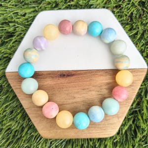 Può includere: Un braccialetto circolare composto da perline rotonde e lisce in una varietà di colori pastello, tra cui rosa tenue, giallo pallido, azzurro e verde menta. Le perline sono disposte in un motivo ripetitivo, creando un accessorio colorato e lucido.
