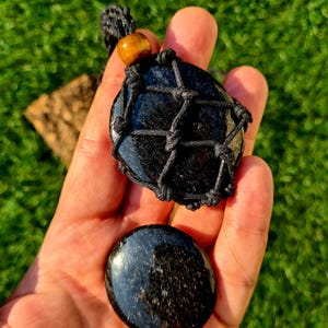 May include: Black obsidian stone pendant with a black macrame cord and a wooden bead. The pendant is in the shape of a circle and is surrounded by a black macrame net. The stone is smooth and polished.