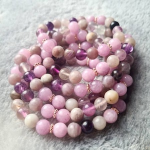 Könnte beinhalten: Ein Stapel rosafarbener und violetter Edelstein-Armbänder mit Roségold-Akzenten. Die Armbänder bestehen aus verschiedenen Steinsorten, darunter Amethyst und Rosenquarz.