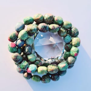 Peut inclure: Un bracelet de perles vertes, rouges et bleues entoure un cristal facetté. Le cristal reflète la lumière, créant un effet scintillant. Le bracelet est un bijou. Les perles mesurent environ 1 cm de diamètre.