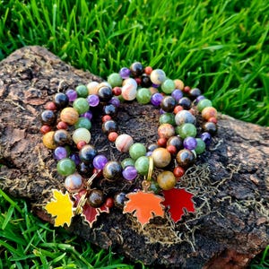 May include: Four bracelets made of various colored stones, including green, brown, purple, and red, with gold leaf accents. The bracelets are stacked on a piece of tree bark and have gold leaf maple leaf charms.