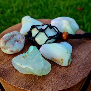 Puede incluir: Una colección de cinco piedras de ópalo verde y azul, algunas con marcas marrones, sobre una superficie de madera. Una piedra está en una bolsa de macramé negra con una cuenta de madera marrón.