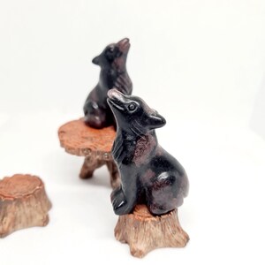 Könnte beinhalten: Zwei dunkelbraune Wolfsfiguren mit rötlichem Farbton, eine auf einem kleinen Holztisch und die andere auf einem Holzstumpf. Die Wölfe sind so geformt, dass ihre Köpfe nach oben geneigt sind. Ein dritter Holzstumpf ist ebenfalls sichtbar.