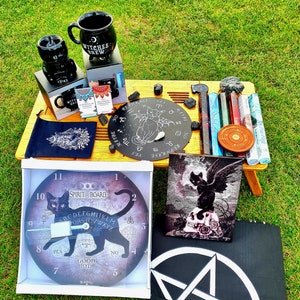 May include: A black and white spirit board with a black cat design, a black and white Ouija board with a black cat design, a black and white pentagram, a black and white cat skull print, a black ceramic mug with the text "Witches Brew", a black ceramic mug with a cat design, a black pouch with a white design, a black and white circular board with a black cat design, a set of incense sticks, and a wooden table.