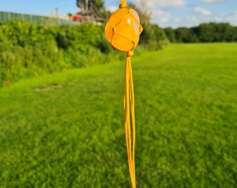 May include: A yellow gemstone pendant with a knotted macrame cord. The pendant is hanging from a yellow cord against a blurred background of green grass and a blue sky.