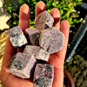 May include: A collection of seven purple and black rough cut gemstones. The stones are in various shapes and sizes, and they are all tumbled and polished.