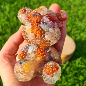 Könnte beinhalten: Eine Kristall-Teddybär-Figur aus klarem Quarz mit Einschlüssen von Amethyst, Citrin und rotem Jaspis. Der Bär sitzt mit ausgestreckten Pfoten.