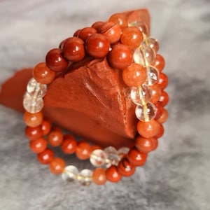 Op de afbeelding: Twee armbanden gemaakt van oranje en heldere kralen. De armbanden liggen op elkaar gestapeld op een stuk oranje-bruine steen.